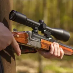 Dłoń trzyma elegancką broń półautomatyczną z lunetą, gotową do polowania w lesie.