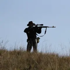 Sylwetka mężczyzny w kapeluszu, celującego z karabinu z lunetą, niczym John Browning, na tle błękitnego nieba.