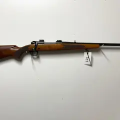 Klasyczny karabin Winchester Model 70 z drewnianą osadą i czarną lufą, zawieszony na białej ścianie.