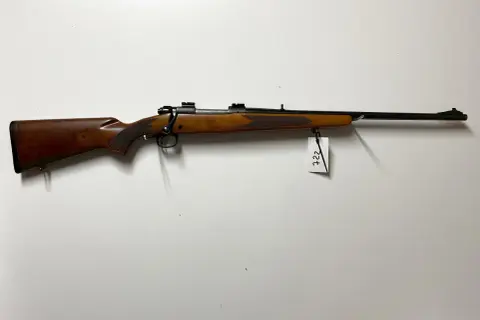 Klasyczny karabin Winchester Model 70 z drewnianą osadą i czarną lufą, zawieszony na białej ścianie.