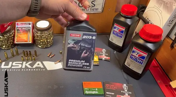 Ręka trzyma butelkę prochu bezdymnego Norma 203-B, obok są spłonki do amunicji śrutowej i inne materiały do elaboracji.