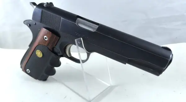 Klasyczny pistolet Colt Government Model kaliber 45, z drewnianą okładziną rękojeści, prezentowany na przezroczystym stojaku.