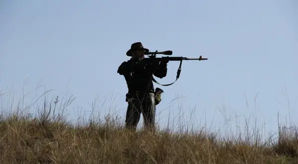 Sylwetka mężczyzny w kapeluszu, celującego z karabinu z lunetą, niczym John Browning, na tle błękitnego nieba.