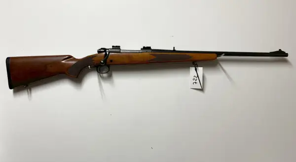 Klasyczny karabin Winchester Model 70 z drewnianą osadą i czarną lufą, zawieszony na białej ścianie.