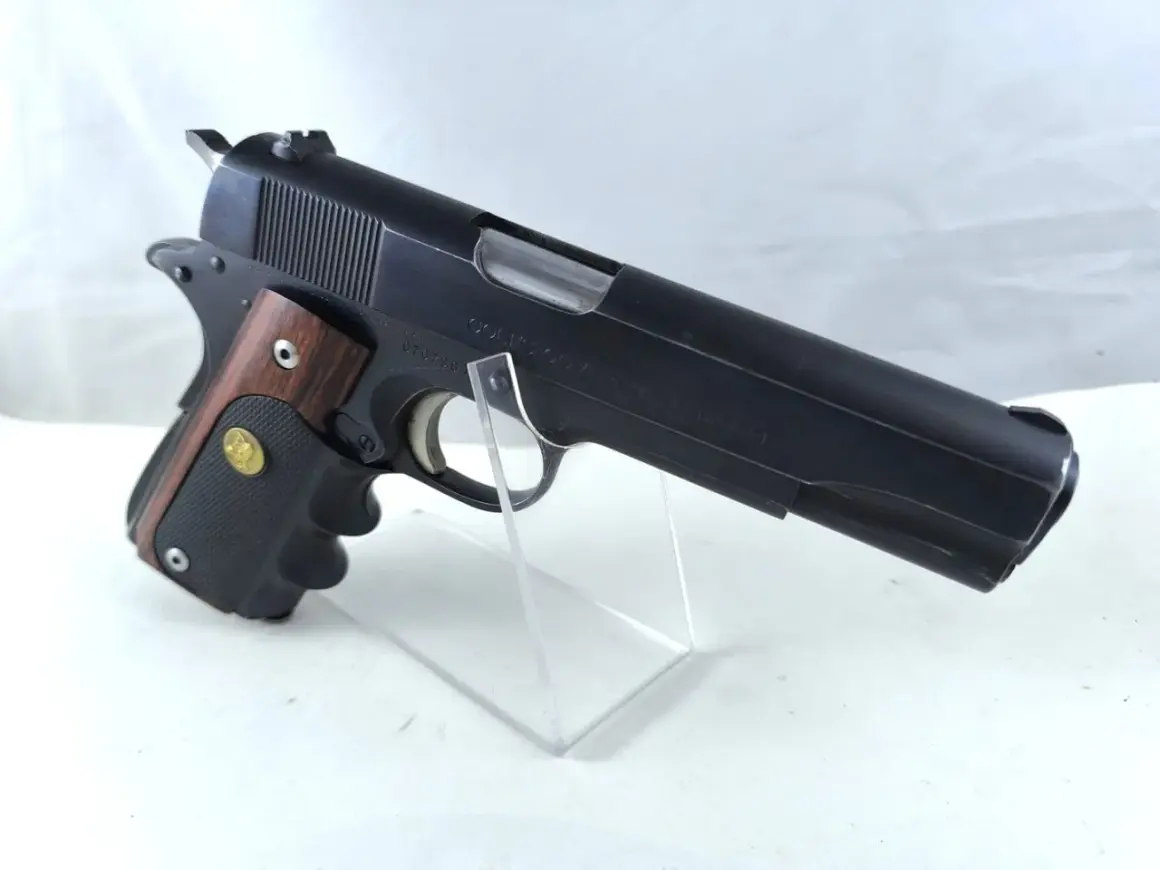 Klasyczny pistolet Colt Government Model kaliber 45, z drewnianą okładziną rękojeści, prezentowany na przezroczystym stojaku.