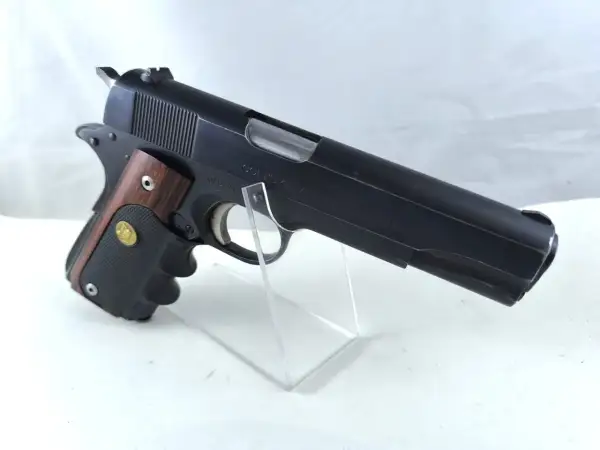 Klasyczny pistolet Colt Government Model kaliber 45, z drewnianą okładziną rękojeści, prezentowany na przezroczystym stojaku.