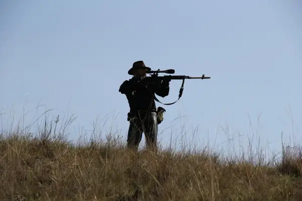 Sylwetka mężczyzny w kapeluszu, celującego z karabinu z lunetą, niczym John Browning, na tle błękitnego nieba.