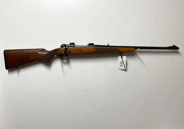 Klasyczny karabin Winchester Model 70 z drewnianą osadą i czarną lufą, zawieszony na białej ścianie.